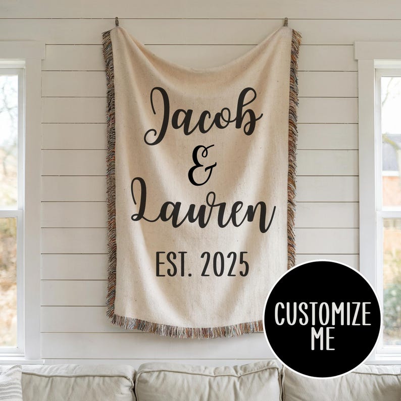 May include: A cream-colored woven throw blanket with a fringe border. The blanket has black text that reads "Jacob & Lauren EST. 2025".