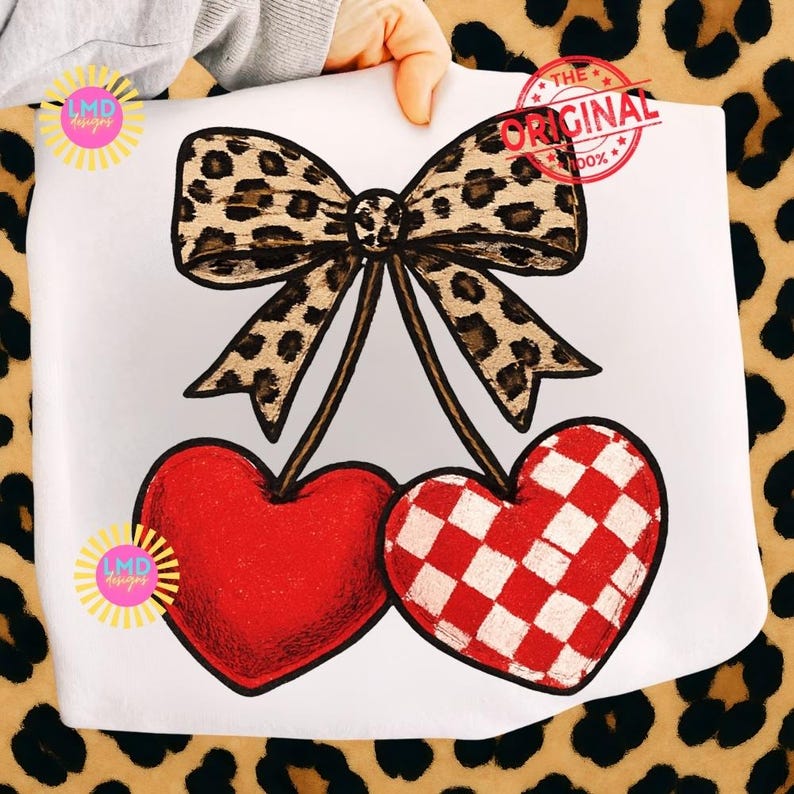 May include: A white print featuring a leopard print bow above two heart-shaped cherries. One cherry is solid red, the other is red and white checkered. The text "THE ORIGINAL" is stamped on the top right.