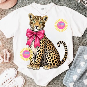 May include: White t-shirt featuring a seated leopard illustration with a large pink bow. The leopard has a spotted coat and green eyes. The shirt also has two sunburst designs with the text "LMD designs" in pink.