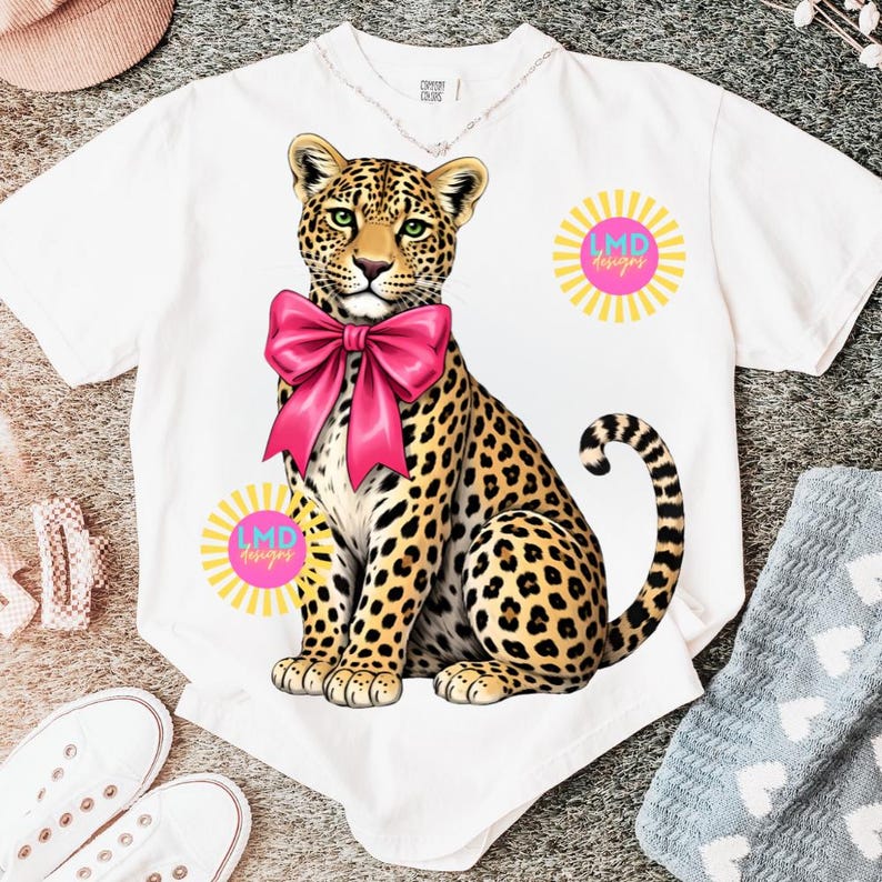 May include: White t-shirt featuring a seated leopard illustration with a large pink bow. The leopard has a spotted coat and green eyes. The shirt also has two sunburst designs with the text "LMD designs" in pink.