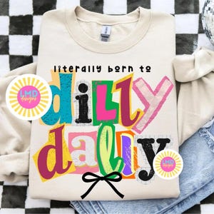 May include: Cream-colored sweatshirt with the text "literally born to dilly dally" in colorful, layered letters. A black bow is at the bottom. The design includes a circular sticker with the text "LMD designs".