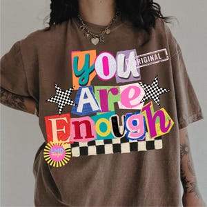 Pu&ograve; includere: T-shirt marrone con un design grafico colorato. Il design presenta le parole "You Are Enough" in uno stile collage con vari colori e caratteri. Include una stella a scacchi e un logo a forma di raggio di sole.