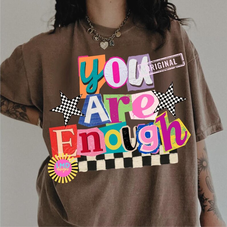 Pu&ograve; includere: T-shirt marrone con un design grafico colorato. Il design presenta le parole "You Are Enough" in uno stile collage con vari colori e caratteri. Include una stella a scacchi e un logo a forma di raggio di sole.