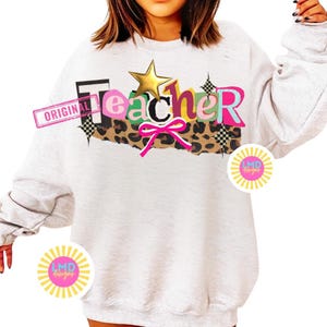 May include: A light gray sweatshirt with the word "Teacher" in colorful, layered text. The design includes a gold star, leopard print, and a pink bow. The word "ORIGINAL" is stamped on the left side of the design.