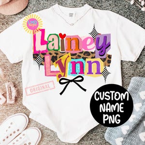 May include: White t-shirt with the name "Lainey Lynn" in colorful, layered text. The design includes a leopard print background, a black bow, and a black circle with "CUSTOM NAME PNG" text. A small "ORIGINAL" stamp is visible.