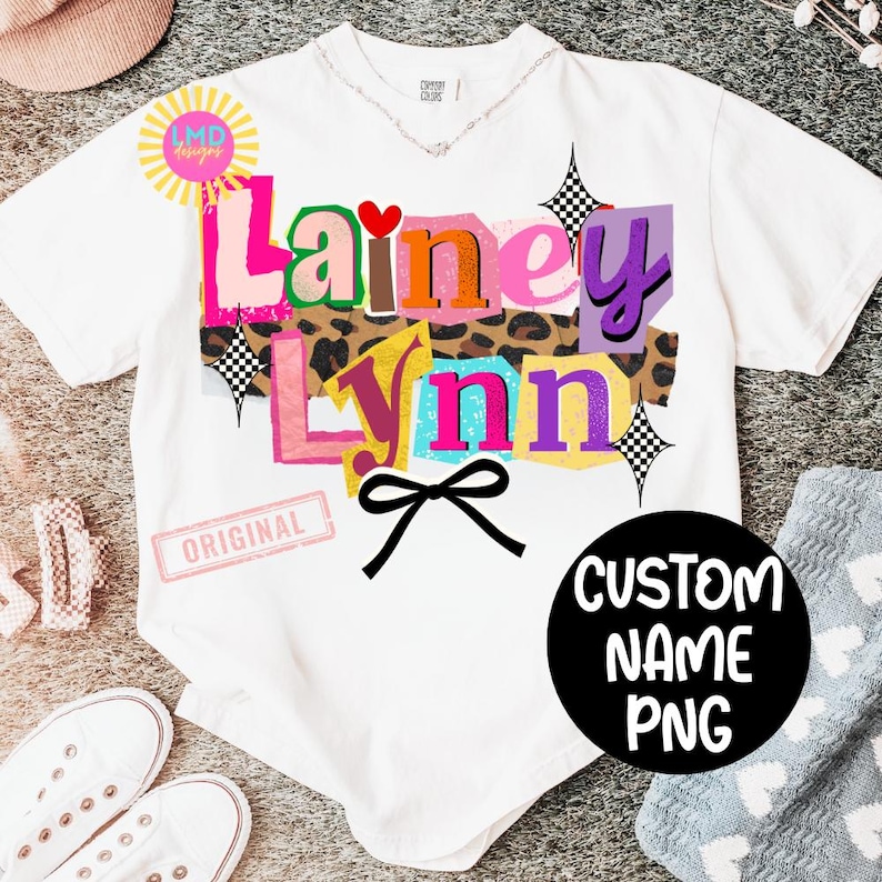 May include: White t-shirt with the name "Lainey Lynn" in colorful, layered text. The design includes a leopard print background, a black bow, and a black circle with "CUSTOM NAME PNG" text. A small "ORIGINAL" stamp is visible.