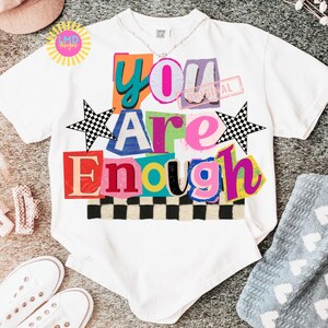 Pu&ograve; includere: T-shirt bianca con la scritta "You Are Enough" a blocchi colorati. Il design include un motivo a scacchiera, stelle e un timbro con la scritta "Original". &Egrave; visibile anche una collana d'argento.