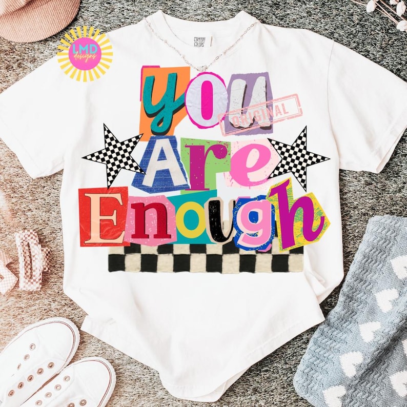 Pu&ograve; includere: T-shirt bianca con la scritta "You Are Enough" a blocchi colorati. Il design include un motivo a scacchiera, stelle e un timbro con la scritta "Original". &Egrave; visibile anche una collana d'argento.