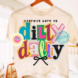 May include: Cream-colored t-shirt with the phrase "literally born to dilly dally" in colorful, blocky letters. A black bow is at the bottom. The shirt has a round logo on the left and a blue and pink stamp that says "the original".
