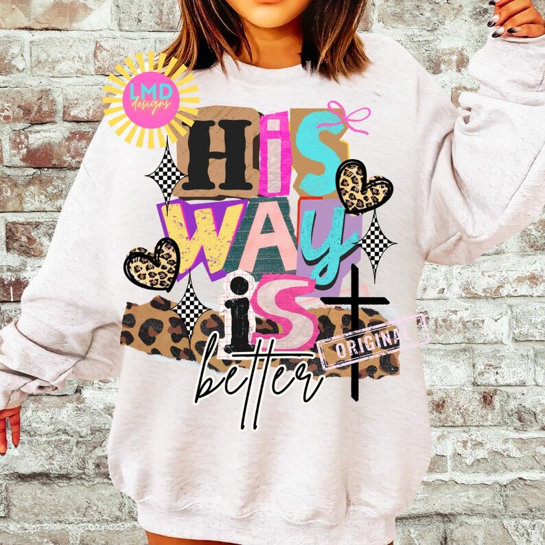 May include: White sweatshirt with the text "His Way Is Better" in a colorful, layered design. The design includes a cross, hearts, and a leopard print pattern. The sweatshirt has long sleeves.