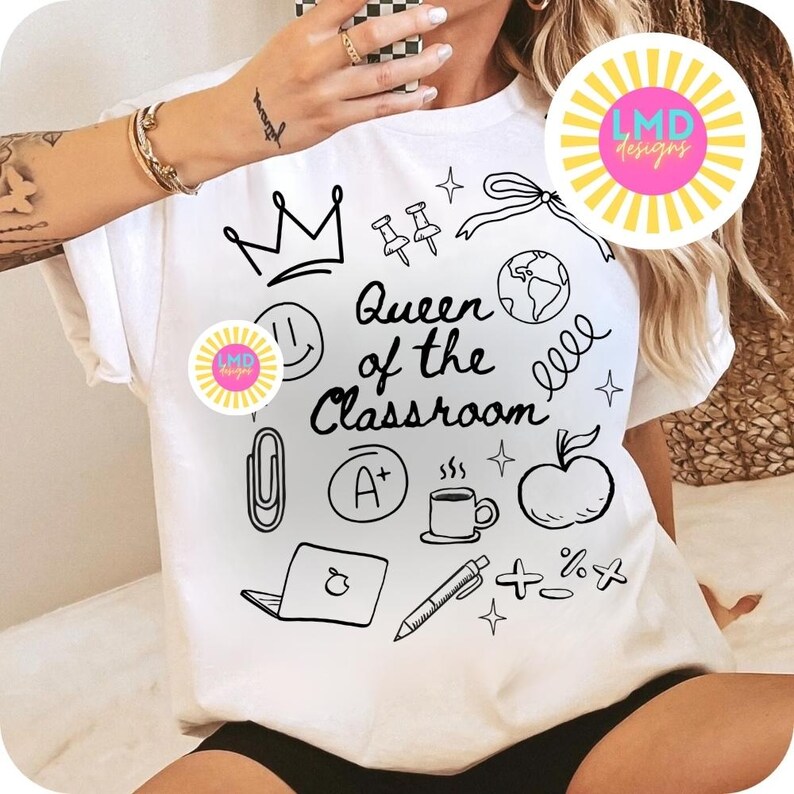 May include: White t-shirt with the phrase "Queen of the Classroom" and various black doodle illustrations, including a crown, apple, and coffee cup. The shirt has a round logo on the front.