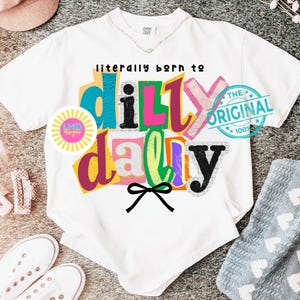 May include: White t-shirt with the text "literally born to dilly dally" in colorful, block letters. A black bow is below the text. The shirt also has a circular logo and a blue stamp that says "The Original 100%."