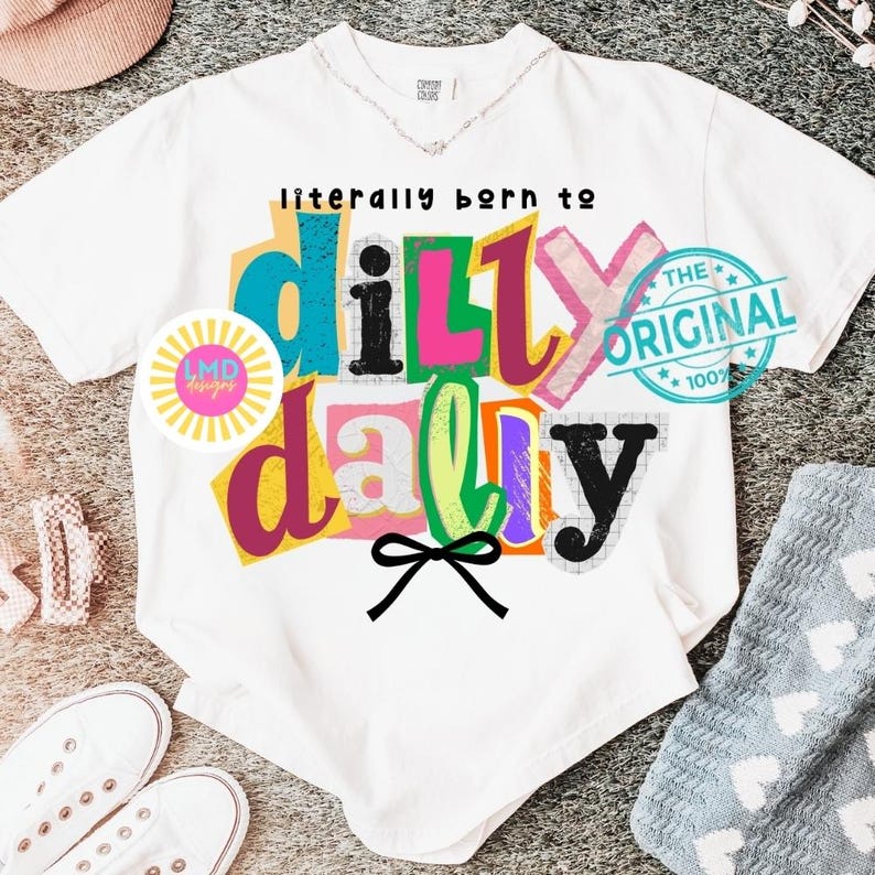May include: White t-shirt with the text "literally born to dilly dally" in colorful, block letters. A black bow is below the text. The shirt also has a circular logo and a blue stamp that says "The Original 100%."