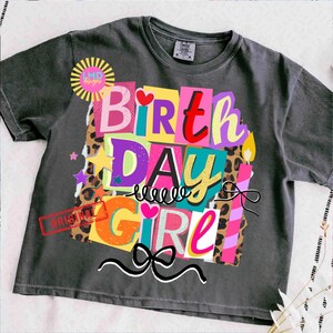 May include: A charcoal gray t-shirt with a colorful graphic that says "BIRTHDAY GIRL." The design features the words in a collage style with various colors, leopard print, stars, and a bow. The shirt has a crew neck and short sleeves.