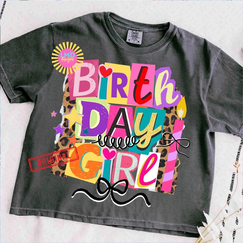 May include: A charcoal gray t-shirt with a colorful graphic that says "BIRTHDAY GIRL." The design features the words in a collage style with various colors, leopard print, stars, and a bow. The shirt has a crew neck and short sleeves.