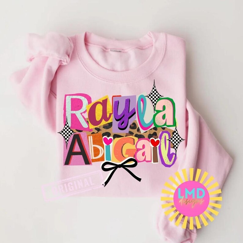 May include: A light pink sweatshirt with the names "Rayla Abigail" in colorful, block letter text. The design includes a leopard print section, hearts, and a black bow. A yellow and pink logo is in the lower right corner.