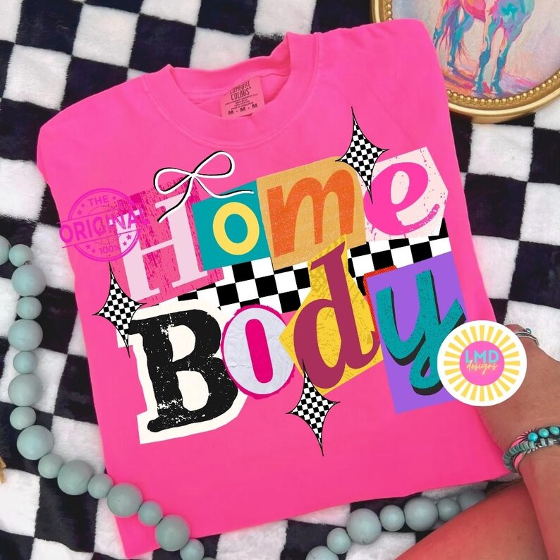 May include: A bright pink t-shirt with the words "Home Body" in a colorful, retro design. The text is layered with various fonts and colors, accented with starbursts and a bow. The shirt is displayed on a black and white checkered surface.