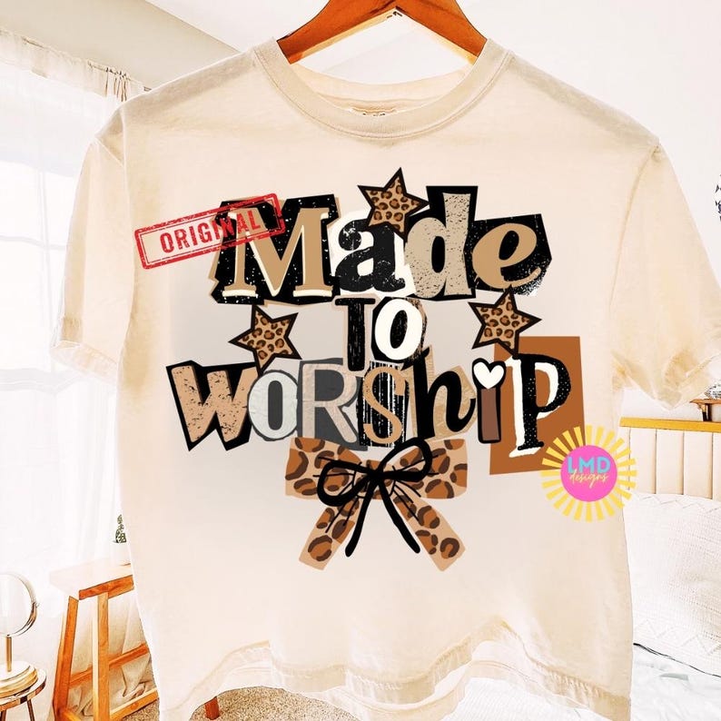 K&ouml;nnte beinhalten: Cremefarbenes T-Shirt mit dem Schriftzug "Made to Worship" in einem mehrschichtigen Design. Das Design umfasst Sterne mit Leopardenmuster und eine Schleife, mit dem Wort "Original" oben links.