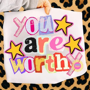 May include: White graphic print featuring the words "YOU ARE WORTHY" in a collage style with various colors and fonts. Yellow stars with black outlines and a small sun logo are also included in the design.