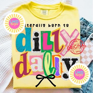 May include: Yellow t-shirt with the phrase "literally born to dilly dally" in colorful, layered text. Features a black bow graphic and two circular logos. The shirt is folded on a denim surface.