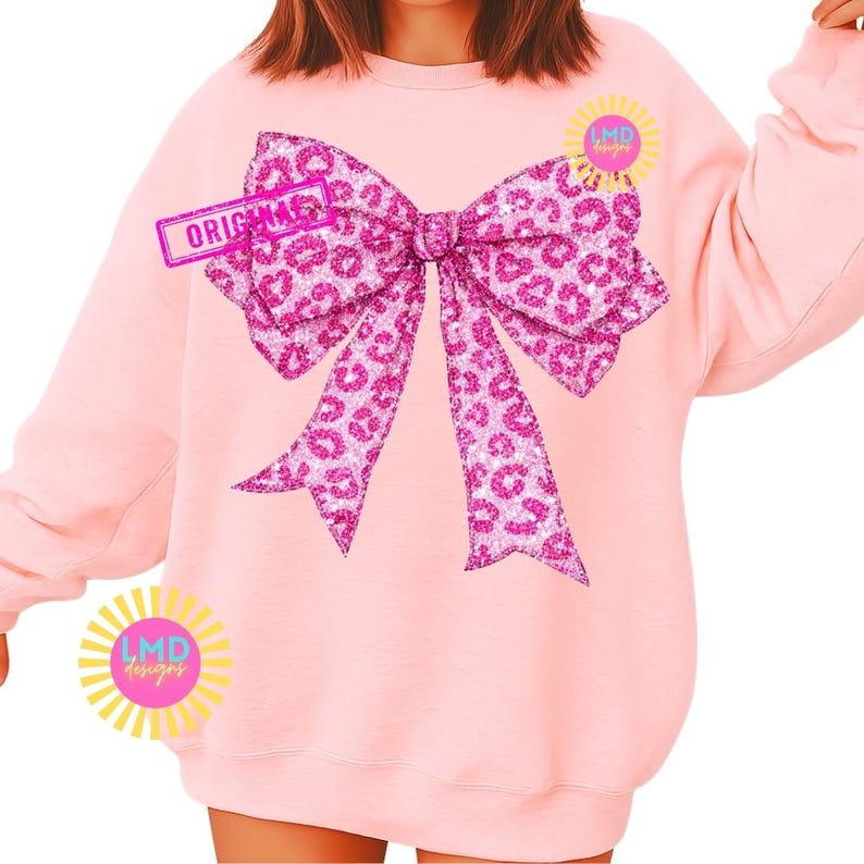 May include: A light pink sweatshirt featuring a large, sparkly pink bow with a leopard print design. The word "ORIGINAL" is printed on a rectangular tag. The sweatshirt also has two circular logos with the text "LMD designs".