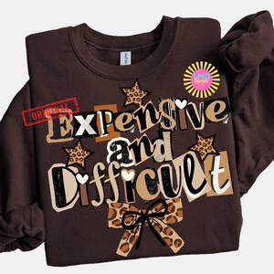 Pu&ograve; includere: Una felpa marrone con le parole "Expensive and Difficult" in un design a strati. Il design include stelle con stampa leopardata, cuori e un fiocco. La parola "Original" &egrave; stampata in rosso.