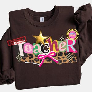 May include: A brown sweatshirt with the word "Teacher" in colorful, layered letters. The design includes a gold star, a pink bow, and a leopard print section. The word "Original" is stamped in red.