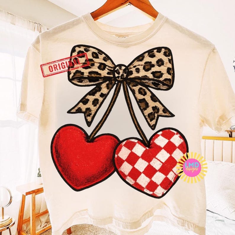 May include: Cream-colored t-shirt featuring a large bow with a leopard print design and two heart-shaped cherries. One cherry is solid red, the other is red and white checkered. The word "ORIGINAL" is stamped on the shirt.
