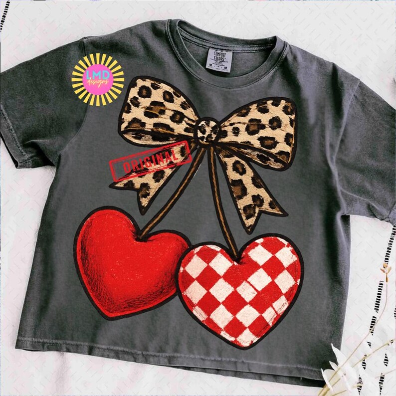 May include: Dark gray t-shirt featuring a leopard print bow and two heart-shaped cherries. One heart is solid red, and the other has a red and white checkered pattern. The word "ORIGINAL" is stamped on a banner.