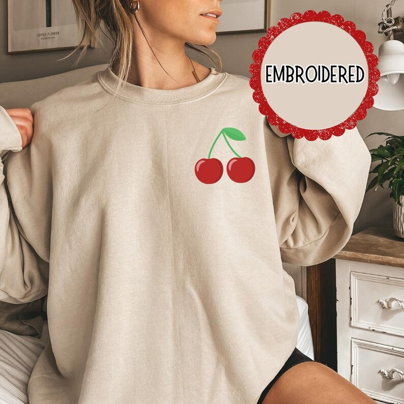 May include: A beige sweatshirt with an embroidered design of two red cherries with green stems on the left chest. The word "EMBROIDERED" is written in a circle on the right side of the image.