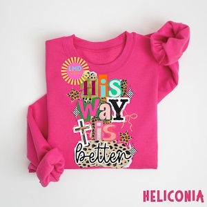 May include: A bright pink sweatshirt with the phrase "His Way Is Better" in a colorful, layered design. The design includes a cross, leopard print accents, and the text "HELICONIA" at the bottom.