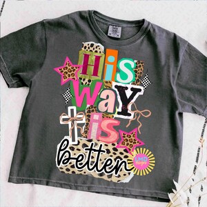 May include: Dark gray t-shirt with the text "His Way Is Better" in a colorful, layered design. The design includes stars, leopard print, and a cross. The shirt has a crew neck and short sleeves.