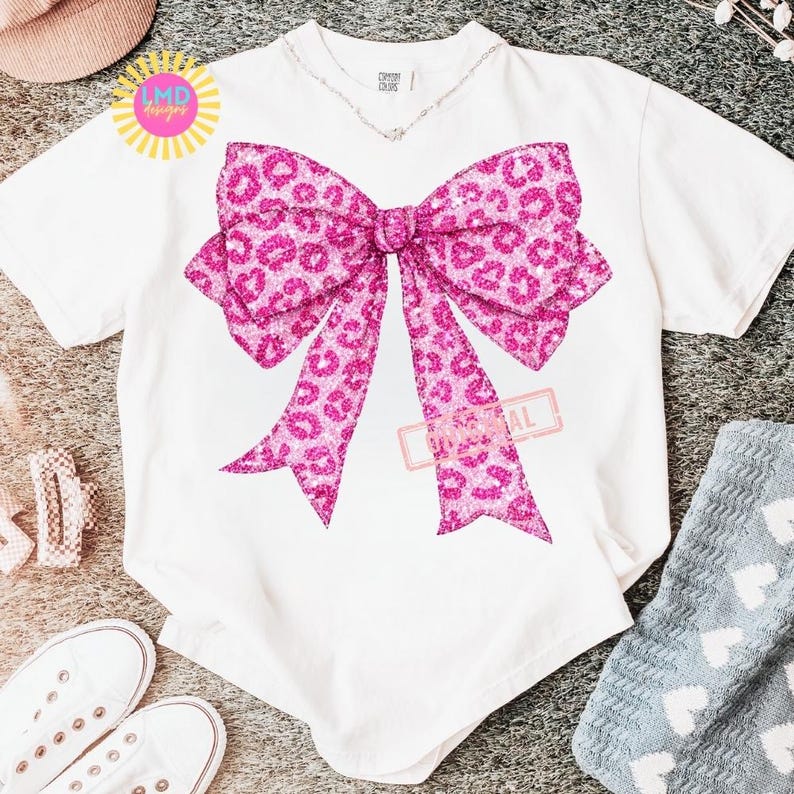 May include: White t-shirt featuring a large pink bow with a leopard print design. The bow is the focal point, with a glittery texture. The shirt has a V-neck and short sleeves. The text "LMD designs" is visible.