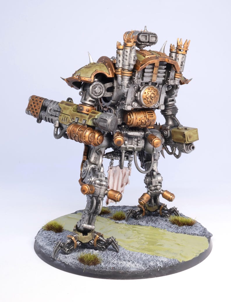 May include: A detailed model of a war machine, featuring a metallic silver body with bronze and olive green accents. The model stands on a textured base with faux grass and a painted path. It has a large weapon and intricate mechanical details.