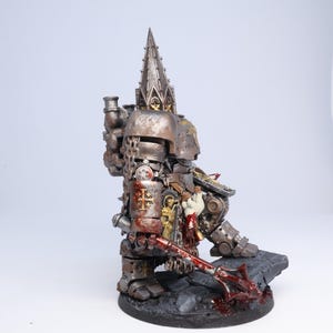 Puede incluir: Figura en miniatura detallada de un guerrero fuertemente blindado, pintada en tonos plateados y bronce met&aacute;licos con detalles en rojo. La figura est&aacute; posada sobre una base texturizada, sosteniendo una maza ensangrentada. La armadura presenta detalles intrincados y una alta aguja ornamentada.