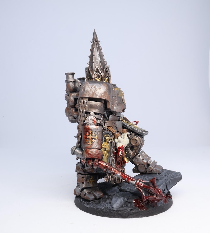 Puede incluir: Figura en miniatura detallada de un guerrero fuertemente blindado, pintada en tonos plateados y bronce met&aacute;licos con detalles en rojo. La figura est&aacute; posada sobre una base texturizada, sosteniendo una maza ensangrentada. La armadura presenta detalles intrincados y una alta aguja ornamentada.