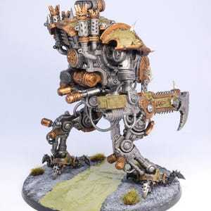 May include: A detailed miniature model of a war machine, featuring a complex design with metallic silver, bronze, and olive green accents. The model stands on a textured base with a painted path and small tufts of grass, showcasing intricate mechanical details.