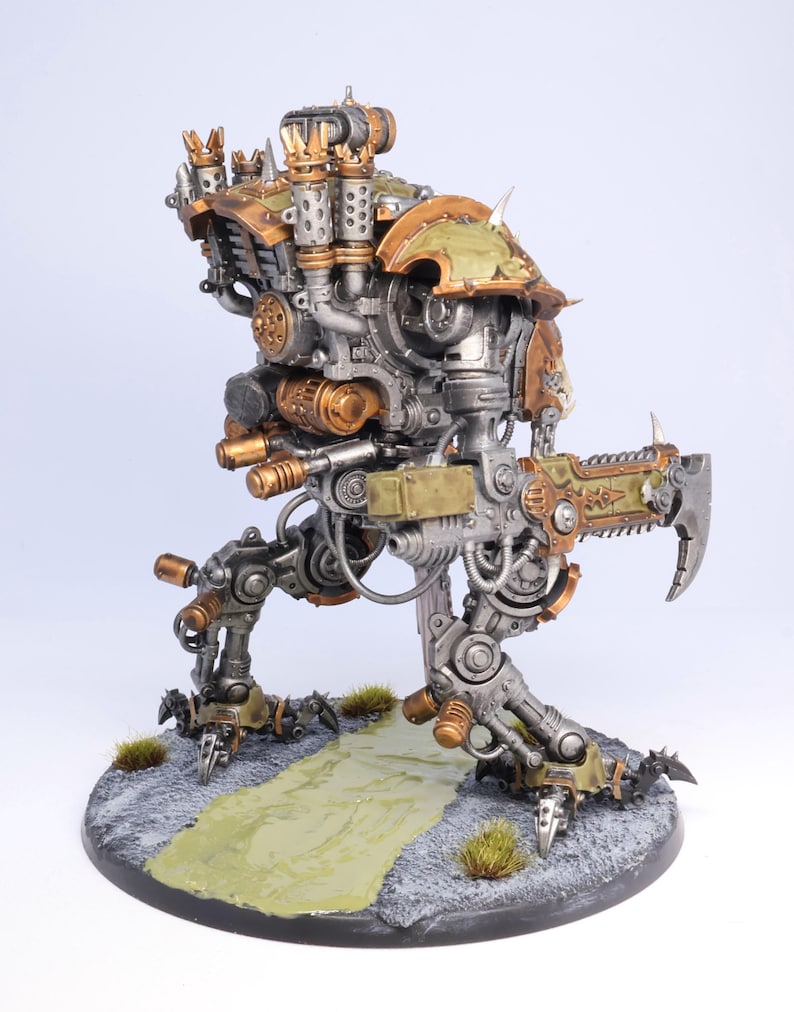 May include: A detailed miniature model of a war machine, featuring a complex design with metallic silver, bronze, and olive green accents. The model stands on a textured base with a painted path and small tufts of grass, showcasing intricate mechanical details.