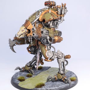 May include: A detailed model of a mechanical war machine, painted in shades of green, bronze, and silver. The model features intricate details, including a skull-like face, sharp claws, and various mechanical components. It stands on a circular base with a painted landscape.