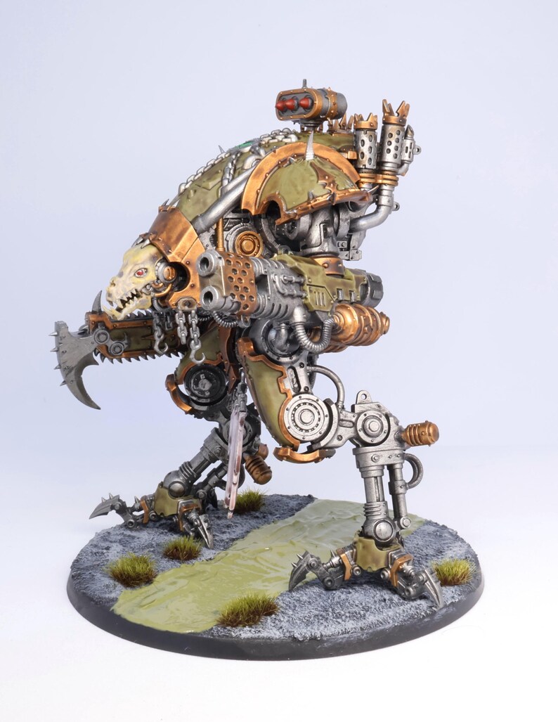 May include: A detailed model of a mechanical war machine, painted in shades of green, bronze, and silver. The model features intricate details, including a skull-like face, sharp claws, and various mechanical components. It stands on a circular base with a painted landscape.