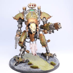 May include: A detailed model of a war machine, featuring a skull-like head and a large gun. The model is painted in shades of green, bronze, and silver, with intricate mechanical details. It stands on a circular base with a painted landscape.