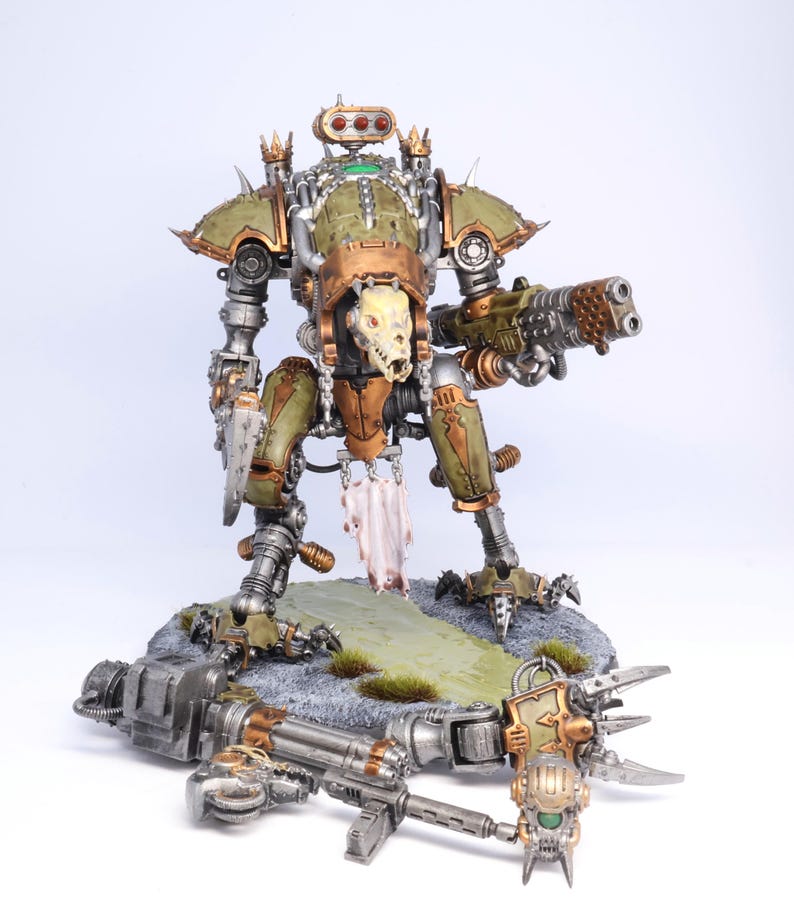 May include: A detailed model of a war machine, featuring a green and bronze color scheme. The model has a skull-like face and is armed with a large gun. It stands on a rocky base with a fallen weapon nearby.