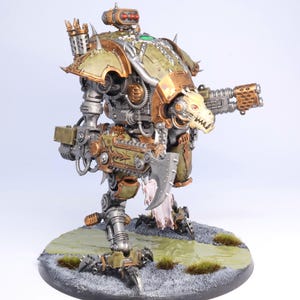 May include: A detailed model of a war machine, featuring a color scheme of olive green, bronze, and silver. The model has a skull-like face on the front and is mounted on a circular base with simulated terrain.