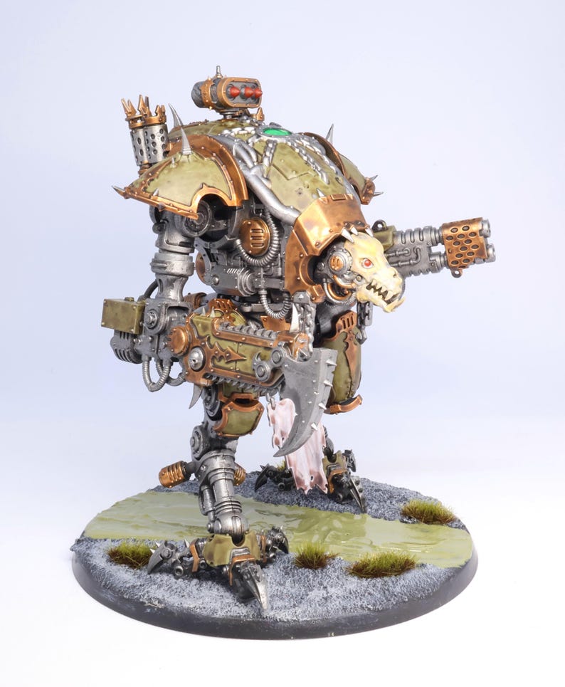 May include: A detailed model of a war machine, featuring a color scheme of olive green, bronze, and silver. The model has a skull-like face on the front and is mounted on a circular base with simulated terrain.
