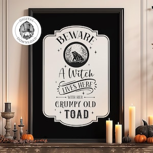 May include: A black and white printable sign with the text "Beware A Witch Lives Here With Her Grumpy Old Toad". The sign is framed and hanging on a wall with a rustic wooden shelf below it. The shelf is decorated with candles, pumpkins, and fall foliage.