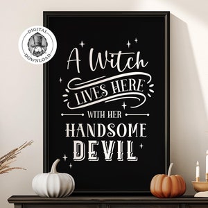Puede incluir: Un cartel imprimible en blanco y negro con el texto "A Witch Lives Here With Her Handsome Devil". El cartel está enmarcado y colgado en una pared sobre una cómoda marrón oscuro con tres cajones. Hay dos calabazas y un candelero con tres velas blancas sobre la cómoda.