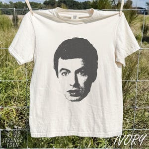 May include: An off-white short-sleeve t-shirt featuring a dark gray graphic print of a person's face centered on the chest. The shirt has a crew neckline and a tag at the inner collar that reads Comfort Colors.