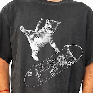 May include: Dark gray t-shirt featuring a white graphic of a tabby cat mid-air, balancing on a skateboard. The skateboard has various stickers and the word "olobe" printed on it. The cat is in a playful pose.