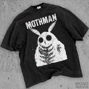 Może przedstawiać: Czarny t-shirt z białym nadrukiem dużej, przypominającej sowę istoty z dużymi oczami i napisem "MOTHMAN" powyżej. Projekt ma postarzany, vintage wygląd. T-shirt ma okrągły dekolt.