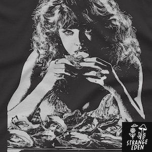 May include: A black t-shirt featuring a high-contrast white graphic print of a person with wavy hair eating an oyster. The person wears rings on their fingers. In the bottom right corner, a small square logo displays a flower, mushroom, and the text STRANGE EDEN.